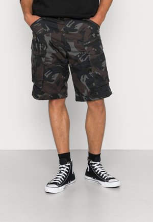 Mężczyzna ubrany w czarne i szare spodenki moro typu cargo oraz czarne sneakersy za kostkę z białymi sznurówkami, stojący z rękami w kieszeniach.