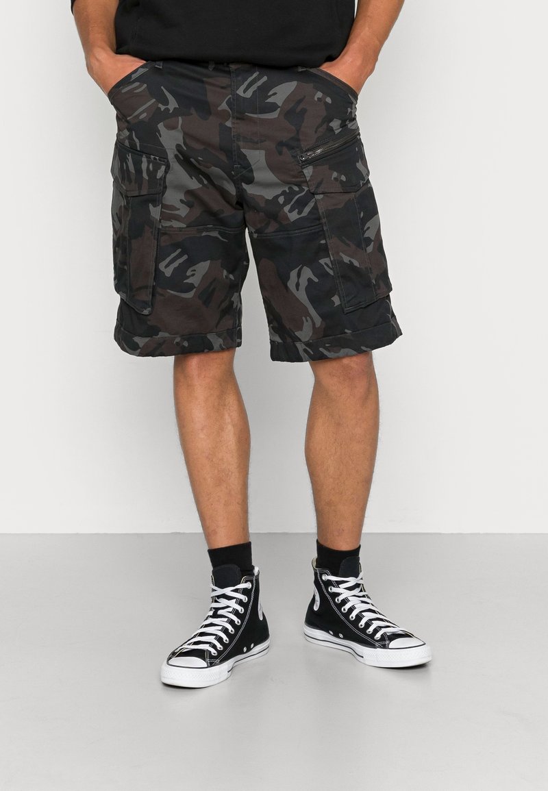 Homme portant un short cargo camouflage noir et gris et des baskets montantes noires avec des lacets blancs, debout les mains dans les poches.