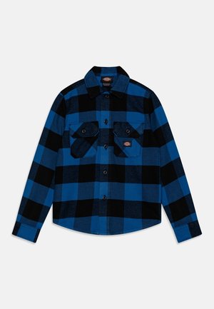 Camicia a quadri blu e nera a maniche lunghe con bottoni e due tasche sul petto con bottone su sfondo bianco.