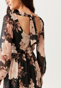 Žena oblečená v černých květovaných šatech s dlouhými průhlednými rukávy a zavázaným odhaleným detailem na zádech, ukazujícími světle růžové a béžové květinové vzory.