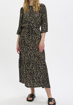 Femme portant une longue robe verte et noire à imprimé léopard avec des manches trois-quarts et des sandales noires à enfiler avec des boucles dorées.