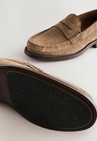 Mocasines de ante marrón con una puntera lisa y redondeada, que presentan una correa decorativa y una suela de goma negra texturizada. Diseño limpio, con acentos mínimos.