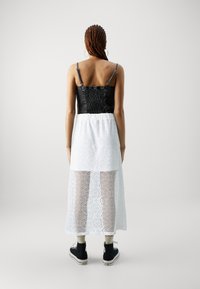 Svart smocket bodice med tynne stropper, som går over til en gjennomsiktig hvit blonder-skjørt med blomster-mønster. Svarte sneakers fullfører stilen.