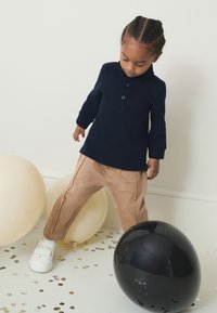 Polo à manches longues bleu marine avec boutons, associé à un pantalon beige avec des coutures latérales. Baskets blanches à scratch. Deux ballons à proximité.