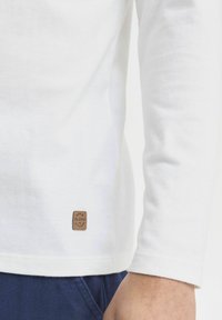 Hvit langermet skjorte laget av mykt stoff med en liten brun lærlabel nederst på kanten. Glatt tekstur, enkelt design.