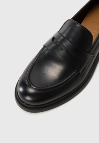 Mocassino in pelle nera con una texture liscia, punta rotonda e una fascia decorativa sulla parte superiore. Suola piatta e dettagli di cucitura minimi.