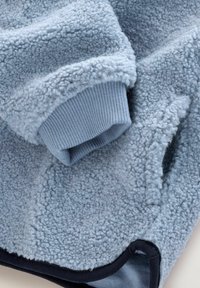 Lichtblauwe fleece stof met een zachte, gestructureerde afwerking. Heeft ribgebreide manchetten in een iets donkerder blauw. Rond afgewerkte randen, marineblauwe bies.