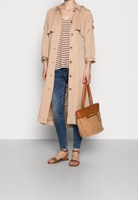 Persona vestida con un abrigo largo beige, una camiseta de rayas, jeans azules, sandalias marrones y sosteniendo un bolso trenzado marrón con acentos de cuero.