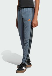 Grå sportbyxor med orange Adidas-logotyp, med svarta sidostriper, elastisk midja och avsmalnande manschetter, matchade med svartvita skor.