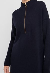 Marinblå stickad klänning med hög krage, prydd med en guldfärgad dragkedja vid halsen. Ribbad textur vid ärmslut och krage, avslappnad passform.