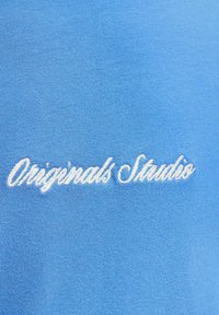 Λευκή κεντητή επιγραφή "Originals Studio" σε μπλε ύφασμα, που εμφανίζει λεπτομερή υφή πλεξίματος.