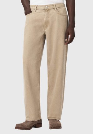 Beige jeans med lige ben båret med brune ruskindsstøvler på en mørkhudet person mod en ensfarvet baggrund.
