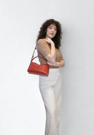 Κόκκινη δερμάτινη τσάντα χειρός με δομημένο σχέδιο, με καμπυλωτή λαβή και υφή φινιρίσματος. Το μοντέλο φοράει μπεζ τοπ και ανοιχτόχρωμο παντελόνι.