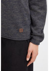 Knitted sweater in dark blue and gray with ribbed cuffs and hem. Features a small brown leather logo patch near the bottom.