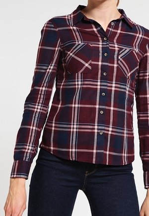 Kvinde iført en langærmet knap-op ternet skjorte i burgundy, navy og hvid med to brystlommer, kombineret med mørkeblå jeans.
