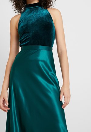 Bezruký tyrkysový sametový top s halterem spojený s tyrkysovou saténovou sukní s vysokým pasem, který nosí osoba s kudrnatými vlasy.