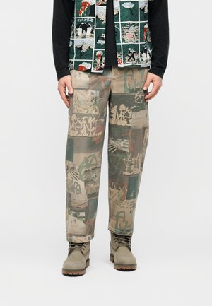 Mann trägt grüne und beige Patchwork-Hose mit Graffiti- und Naturmotiven, kombiniert mit grauen Schnürstiefeln.
