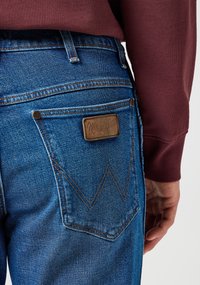 Mėlyni džinsai su ruduo odiniu užsegimu, pažymėtu "Wrangler", ant užpakalinių kišenių, su zigzago siuvinėjimo detalėmis.