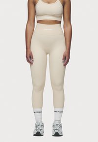 Beige athletic leggings with a high waistband, smooth texture, and logo detailing. Paired with white socks and gray running shoes.