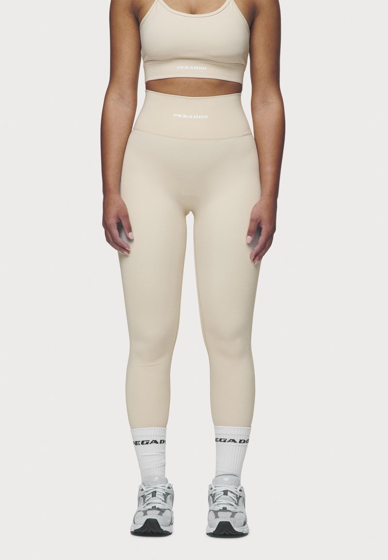 Beige athletic leggings with a high waistband, smooth texture, and logo detailing. Paired with white socks and gray running shoes.