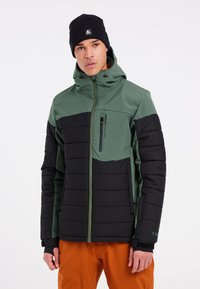 Groene en zwarte pufferjack met een ritsborstzak, verstelbare capuchon, lange mouwen en geribde manchetten, gedragen met oranje broek.