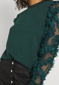 Mujer vistiendo un suéter verde oscuro con mangas transparentes adornadas con aplicaciones florales en 3D y pantalones negros de talle alto con botones plateados.