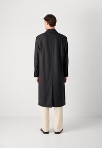Ein Mann, der von hinten steht, trägt einen langen dunklen Hahnentritt-Mantel, helle weite Hosen und schwarze Schuhe vor einem schlichten weißen Hintergrund.