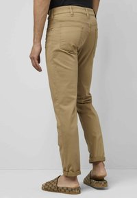 Beige Chinos mit schmaler Passform, ausgestattet mit Gesäßtaschen und dezenten Stickdetails, an den Knöcheln umgeschlagen, kombiniert mit gemusterten Slippern.