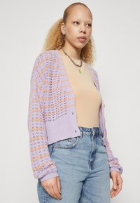 Violeta adīta džemperis ar oranžiem un baltiem rakstiem, ribotām malām un piedurknēm, ar pogām un brīvu piegriezumu. Ikdienas apģērbs virs kailas blūzes.