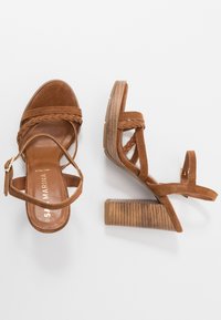 Brown suede sandals with braided straps, open toe, and a chunky wooden heel featuring a textured finish. Adjustable ankle strap included.