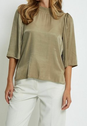 Femme portant un chemisier en satin vert olive avec des manches bouffantes trois quarts et un pantalon blanc taille haute sur un fond uni.