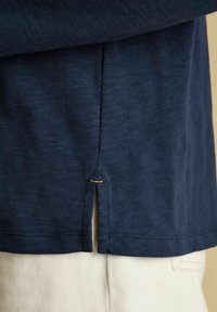 Tumedast sinisest särgi äärest, millel on külgsisene lõhe, kreemivärviliste pükste peal.