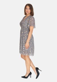 Blumenkleid mit tiefem V-Ausschnitt und kurzen Schmetterlingsärmeln, auf einem marineblauen Hintergrund mit gelben und weißen Mustern. Getragen mit schwarzen Absätzen.