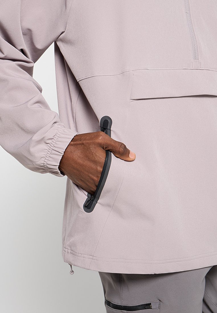 Dunkelhäutige Hand in einer hellrosa Jackentasche mit einem schwarzen Reißverschluss, zeigt elastische Bündchen und einen Teil grauer Hosen mit Reißverschlusstasche.