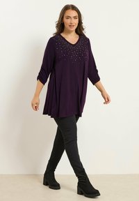 Lila tunika med V-ringning, dekorerad med silvernitar vid halsringningen, tillsammans med svarta skinny jeans och svarta stövlar med medelhöjd.