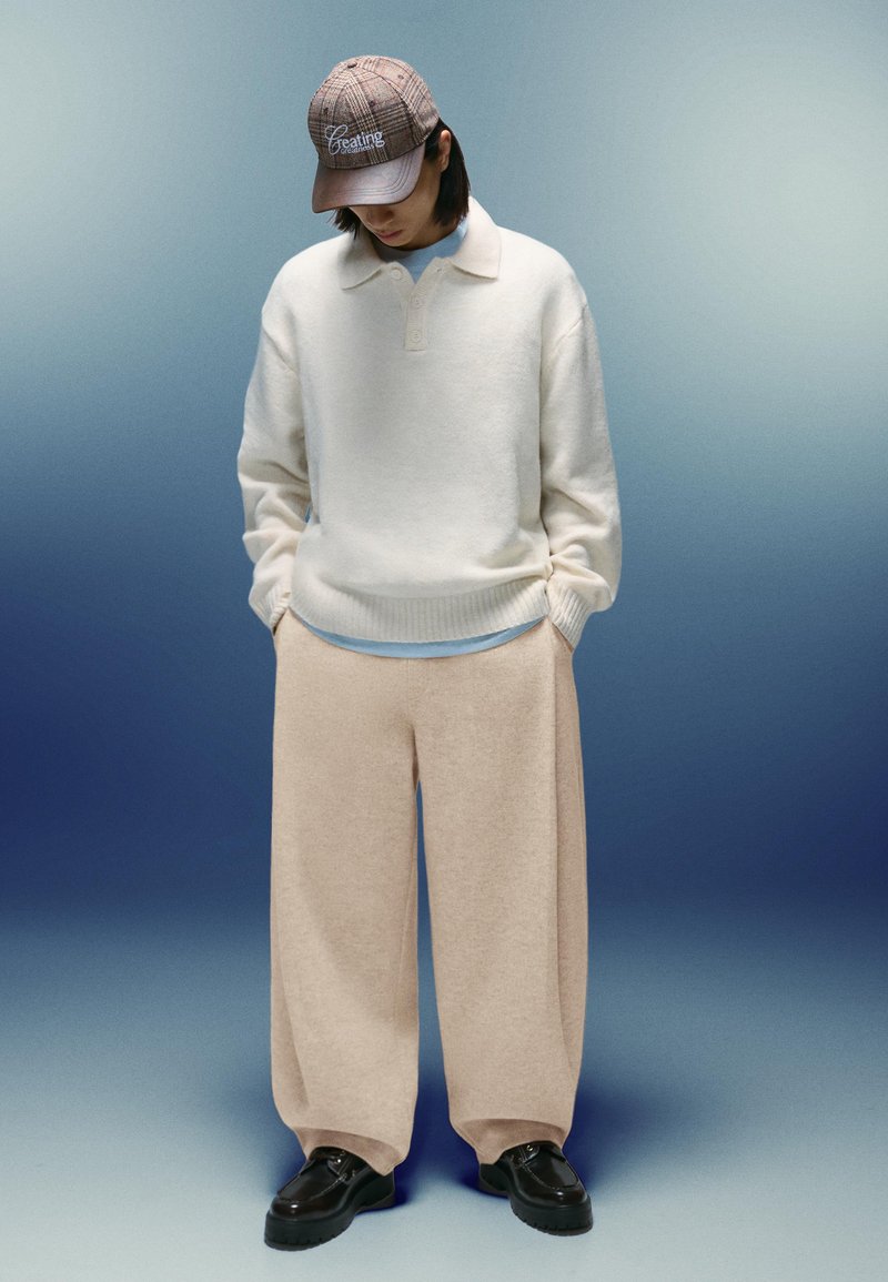 Osoba v béžových širokých nohaviciach, krémovom svetri, hnedom kockovanom klobúku a čiernych topánkach, stojí s rukami v vreckách a s hlavou naklonenou nadol.