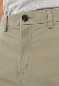 Pantalones verde oliva claro hechos de una tela suave. Presentan un botón negro, lazos para cinturón y una costura sutil a lo largo de las costuras.
