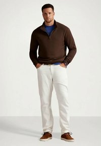 Άνδρας στέκεται με τα χέρια στις τσέπες, φορώντας καφέ πουλόβερ με τρίκοπο, μπλε πουκάμισο, λευκά παντελόνια, καφέ παπούτσια και ένα ρολόι στον αριστερό καρπό.