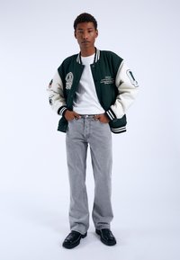 Chaqueta tipo varsity verde y blanca con parches bordados, puños a rayas en blanco y negro, combinada con jeans grises y zapatos negros.