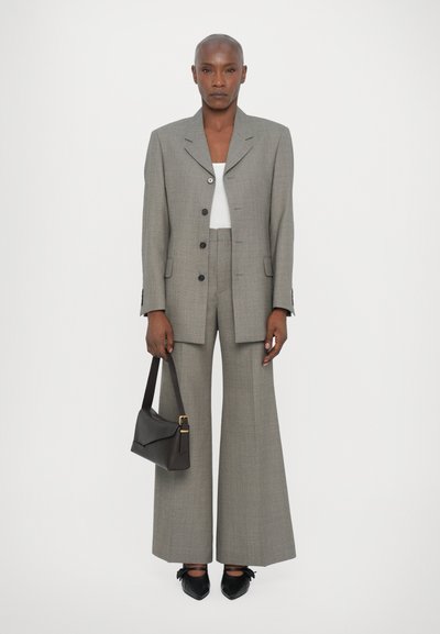 Grijze check blazer met gelapte revers, bijpassende brede broek en een witte top. Houdt een kleine donkerhandtas vast en draagt zwarte puntige schoenen.