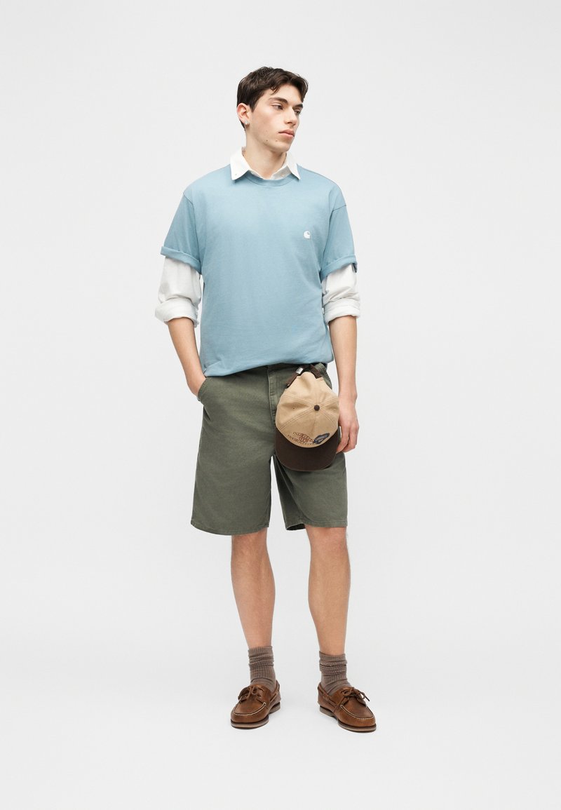 Jeune homme vêtu d'un T-shirt bleu clair par-dessus une chemise blanche, d'un short vert olive, de chaussures marron, tenant une casquette beige et marron, debout devant un fond blanc.