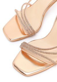 Sandalias doradas de punta abierta con frentes cuadrados y tres delgadas correas brillantes de pedrería a lo largo de la parte superior.