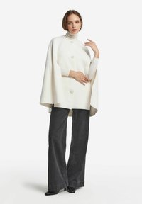 Kobieta ubrana w białą płaszczową pelerynę z guzikami na białym golfie, czarne spodnie o szerokich nogawkach oraz czarne szpilki, stojąca na białym tle.