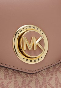 Gros plan sur un sac à main beige Michael Kors montrant le logo MK circulaire doré et les détails de couture sur un matériau texturé.