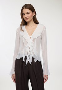 Giovane donna che indossa una camicetta bianca trasparente con volant e maniche lunghe, e pantaloni neri a gamba larga e vita alta, in piedi davanti a uno sfondo neutro.