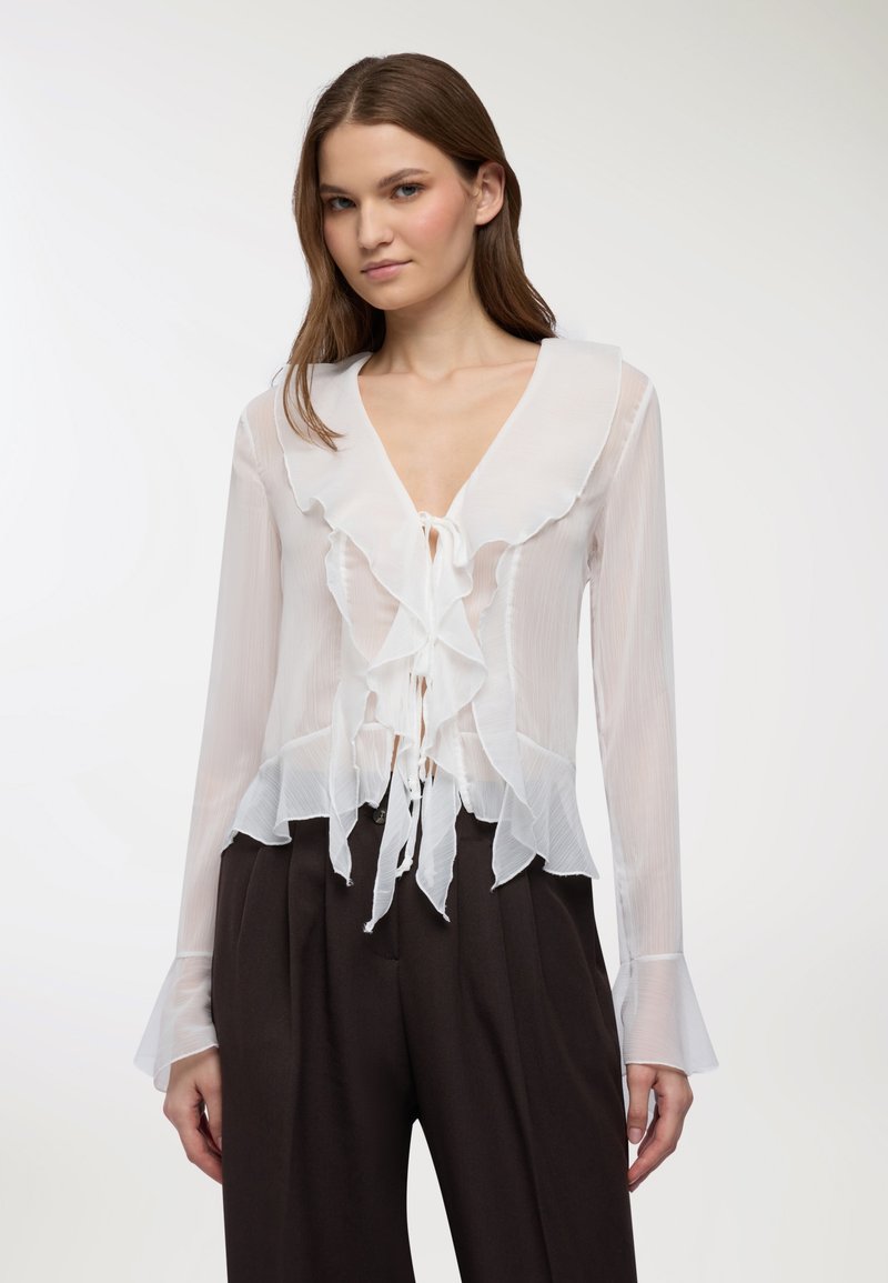 Giovane donna che indossa una camicetta bianca trasparente con volant e maniche lunghe, e pantaloni neri a gamba larga e vita alta, in piedi davanti a uno sfondo neutro.