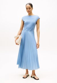Himmelblaues, gefältetes Kleid mit einem enganliegenden, gerippten Oberteil, kurzen Ärmeln und schwingendem Rock. Das Model hält eine cremefarbene Handtasche und trägt schwarze Flip-Flops.