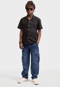 Zwart T-shirt met korte mouwen met een abstract patroon, gecombineerd met blauwe cargobroek en witte sneakers. Accessoires zijn een bruine beanie en een zonnebril.