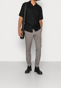 Camisa negra de manga corta con botones, pantalones grises con estampado y botines negros; se lleva un bolso de hombro cruzado.