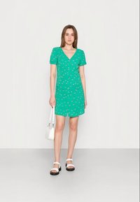 Vestido curto verde com padrão floral, decote em V, mangas curtas e cintura ajustada. Usado com sandálias brancas e uma pequena bolsa branca.